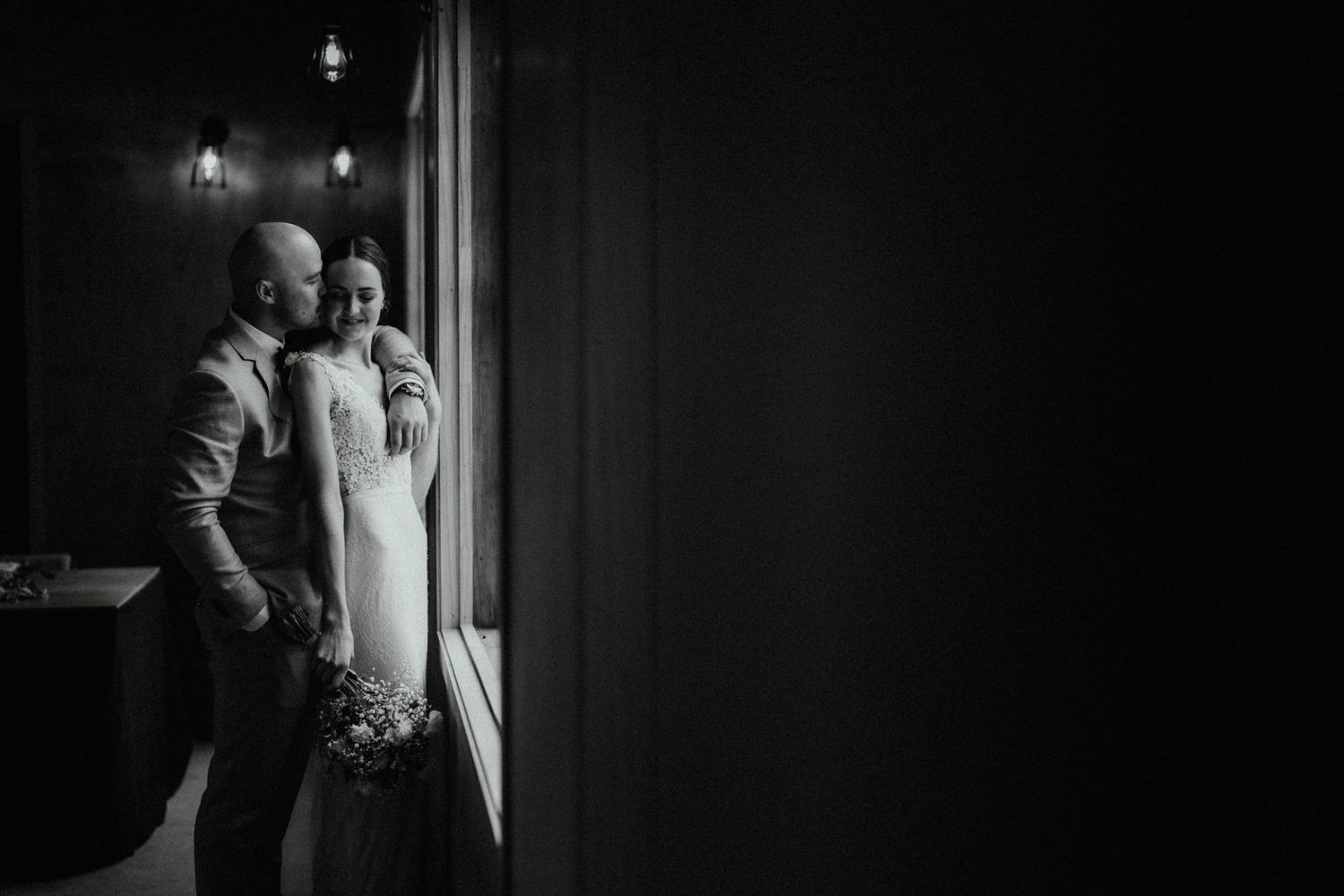 Black and white portrait of a groom embracing his bride from behind beside a tall window, holding a bouquet.