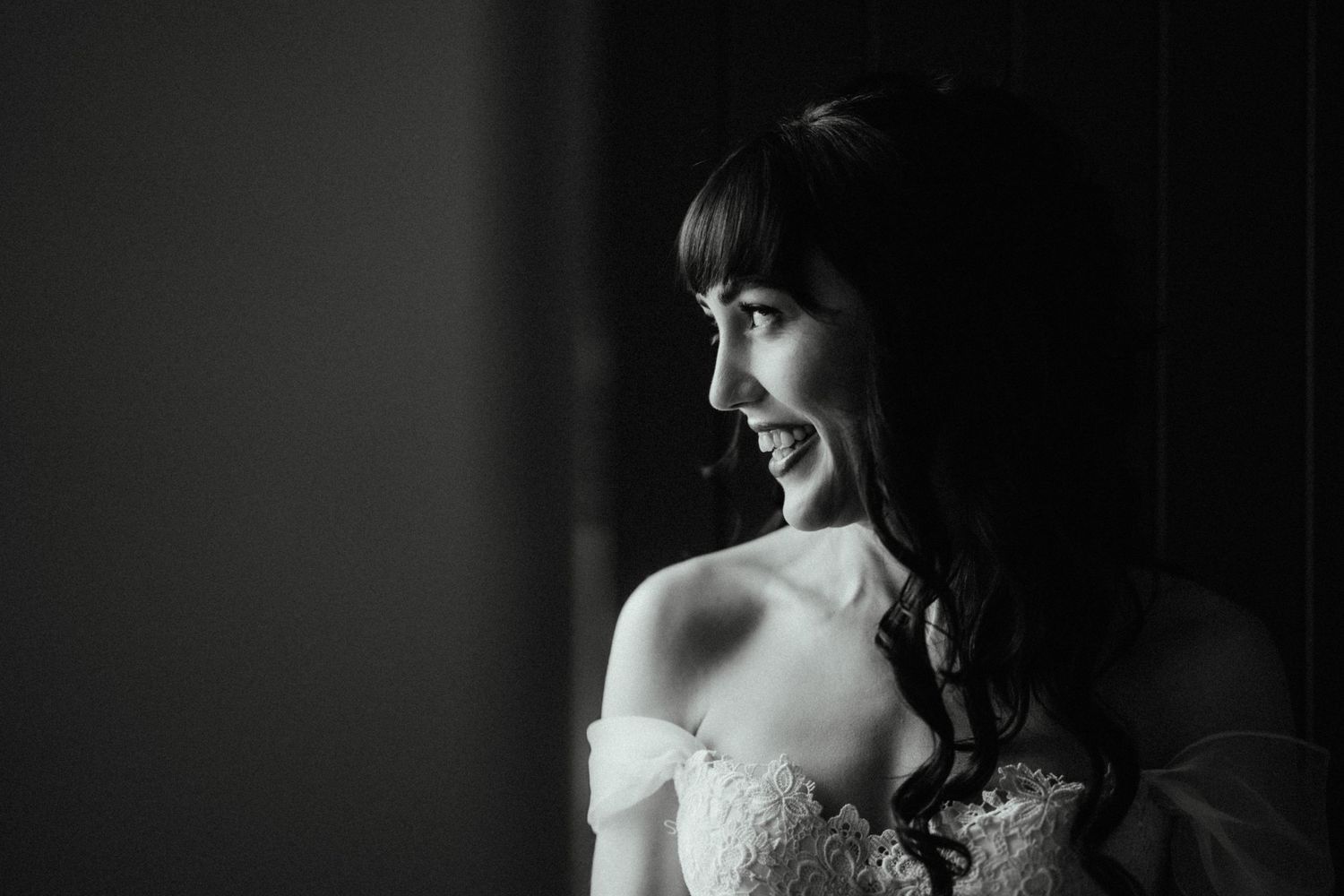 Black and white portrait of a smiling bride with bangs and an off-the-shoulder lace gown gazing toward a window.