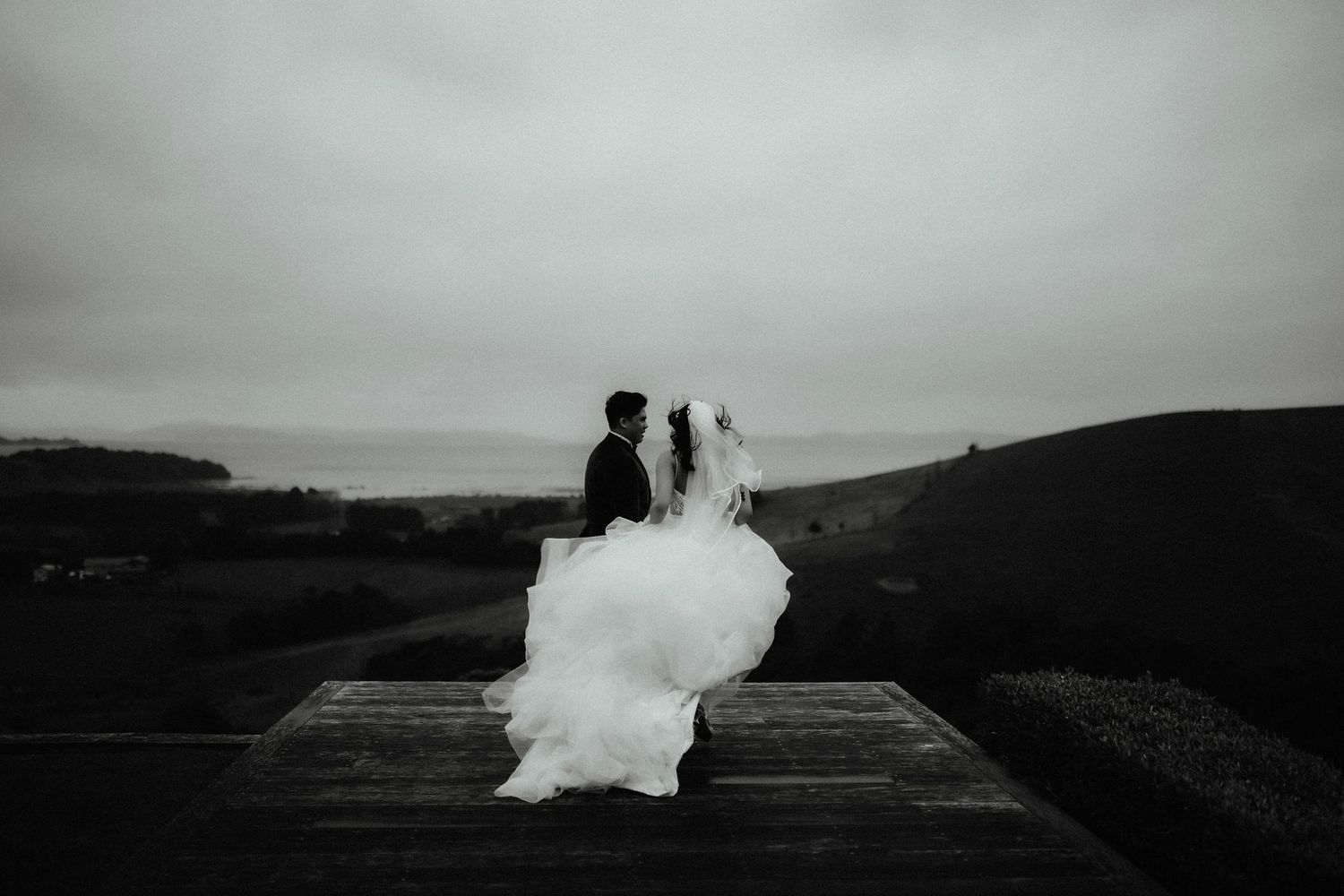 Black and white photo of a bride in a flowing white gown and groom on a wooden deck overlooking a moody coastal landscape.