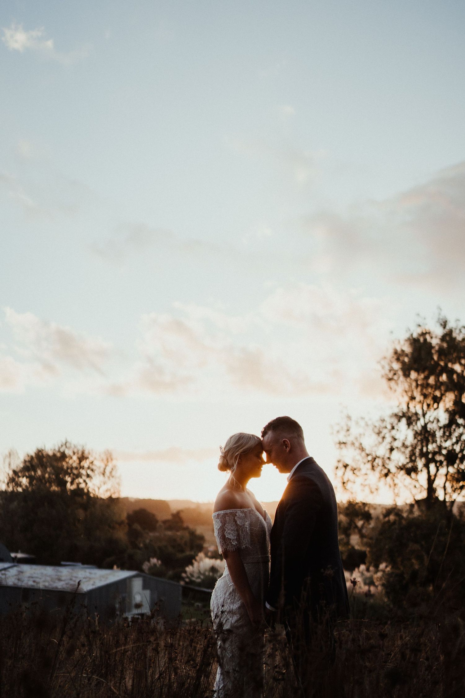 Bride and groom touch foreheads in a quiet moment at sunset with silhouetted trees and soft clouds behind them.