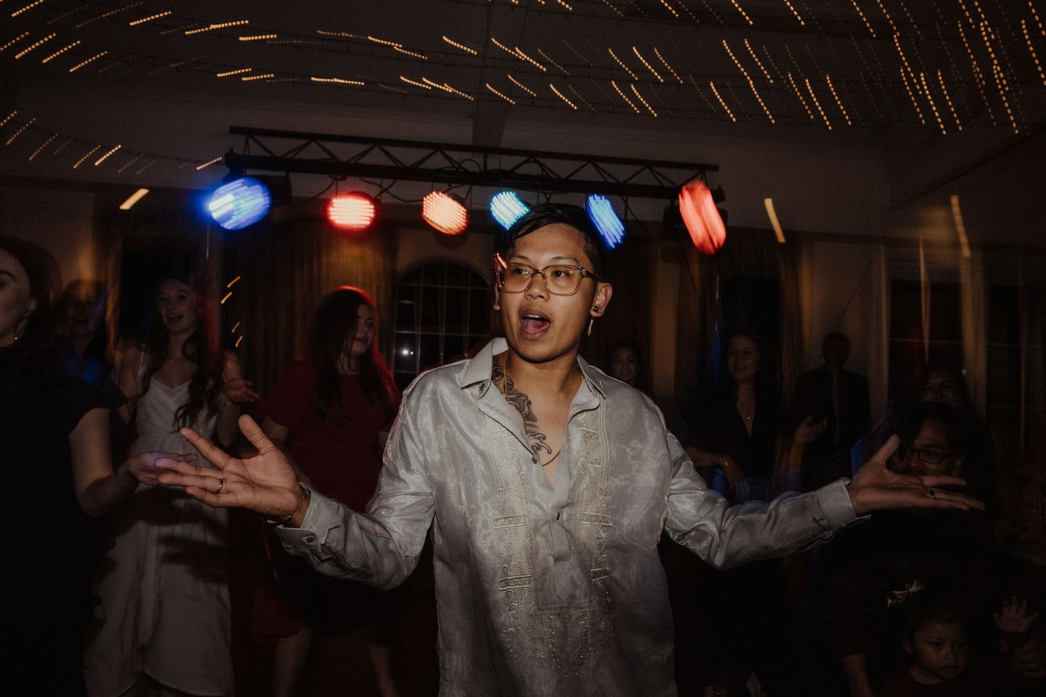 Guest in glasses dances joyfully with arms outstretched at a wedding reception under colorful stage lights.