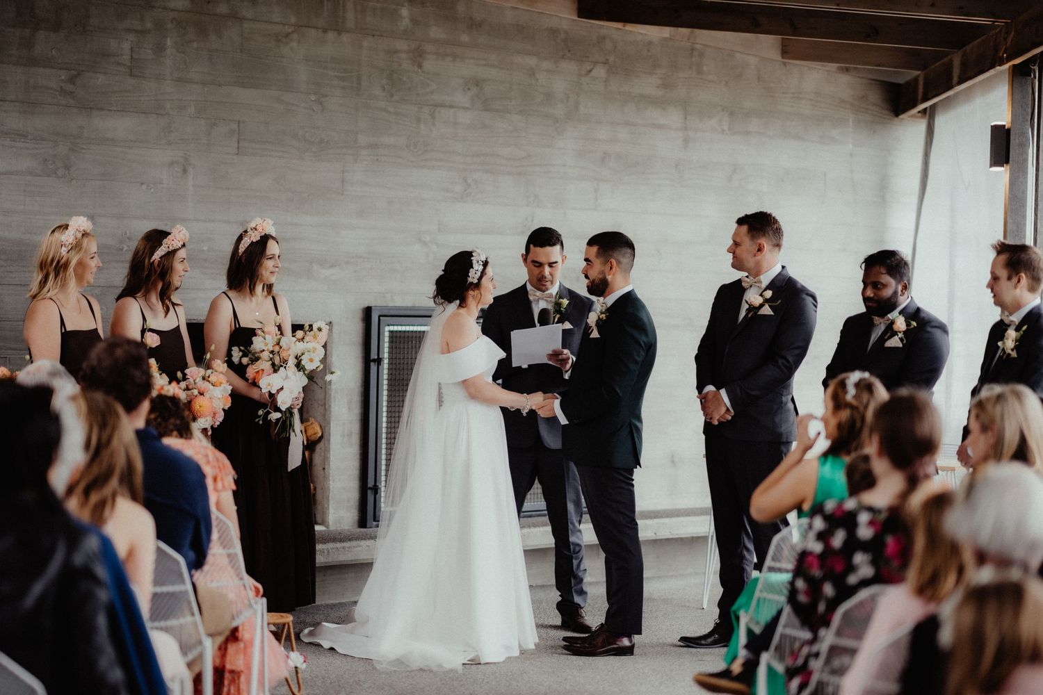 Bride and groom exchange vows before an officiant as groomsmen, bridesmaids and seated guests watch the wedding ceremony.