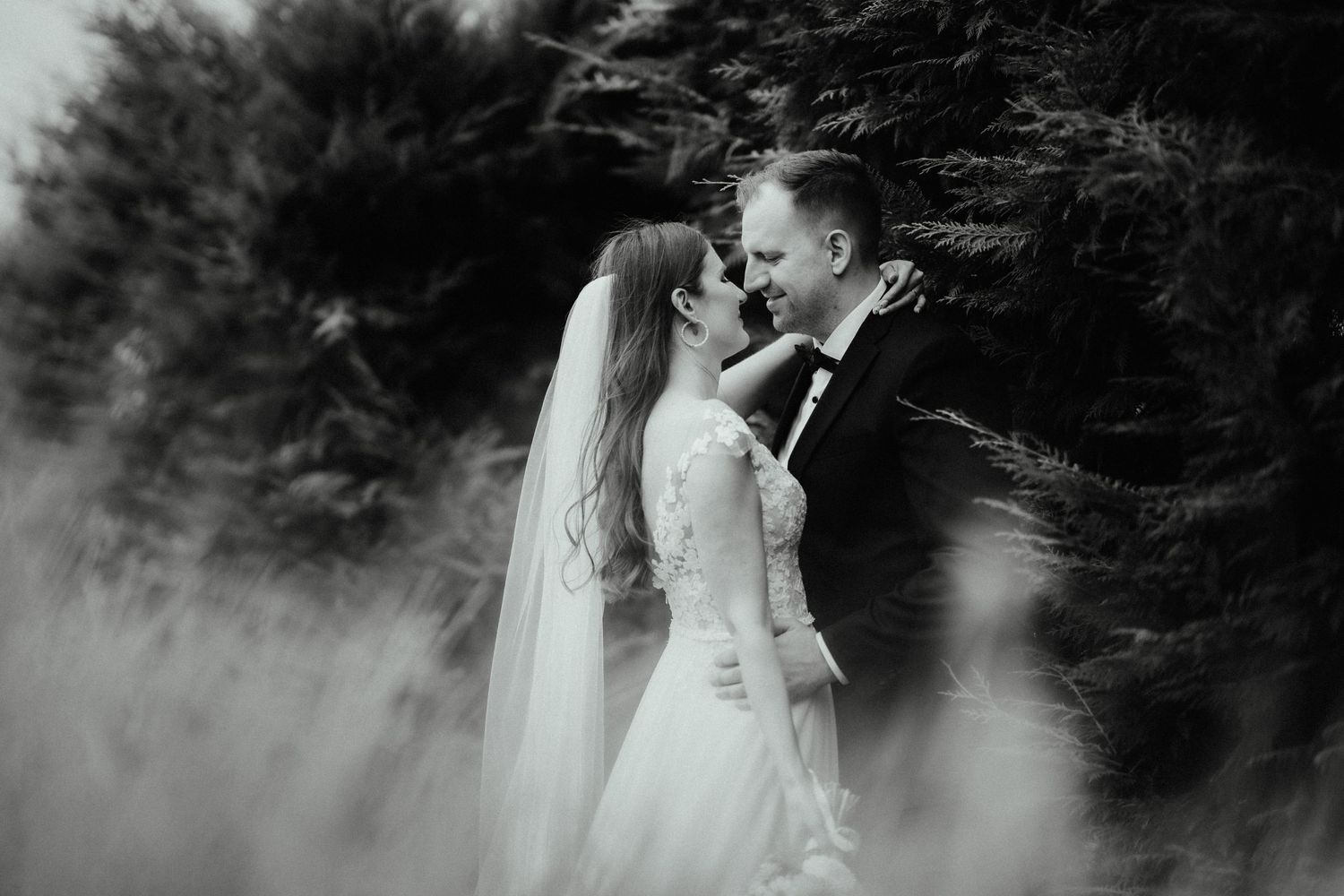 Black and white photo of a bride and groom embracing in a soft-focus field with trees in the background.