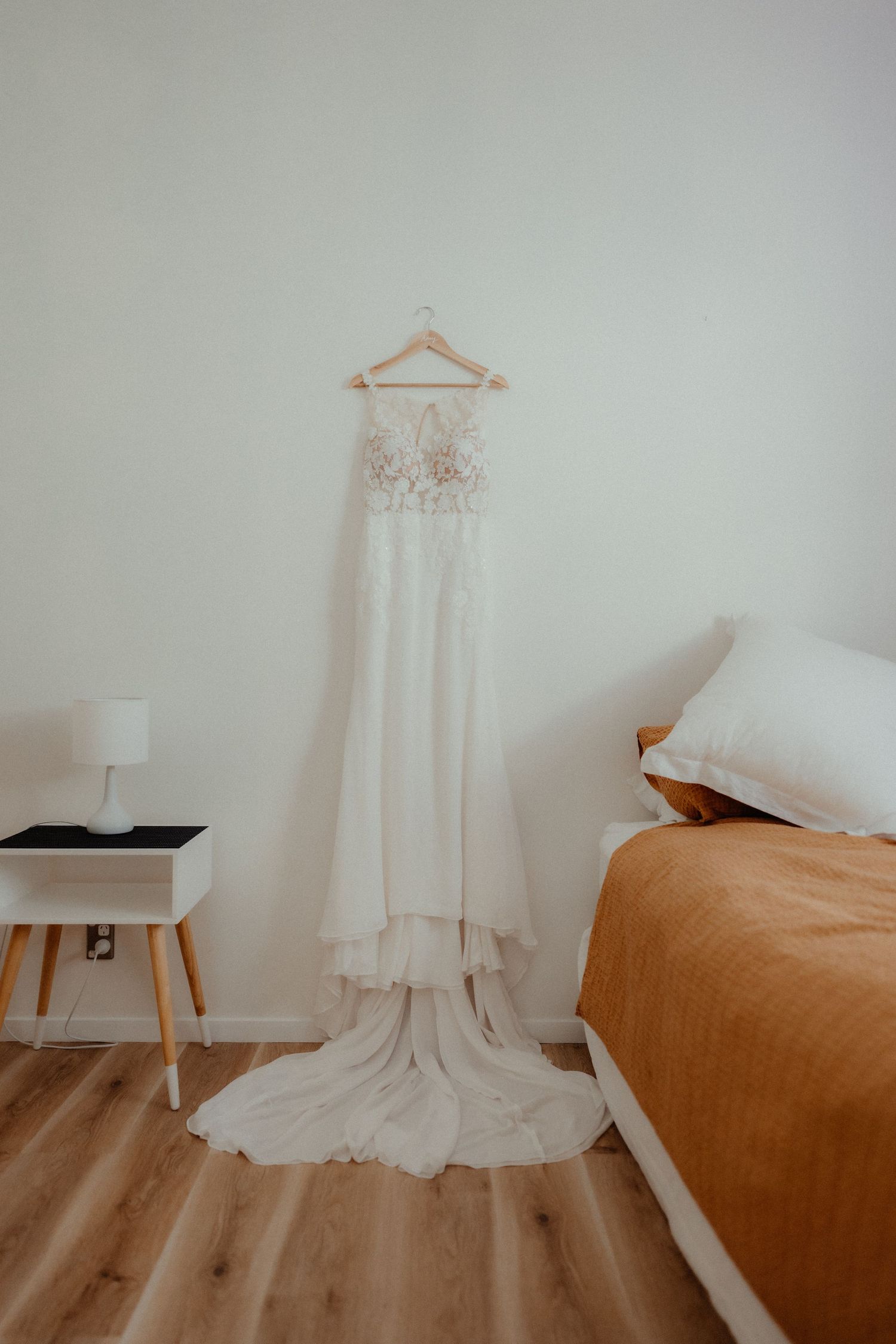 Lace wedding gown with a long train hangs from a wooden hanger on a white wall beside a bed in a bright bedroom.