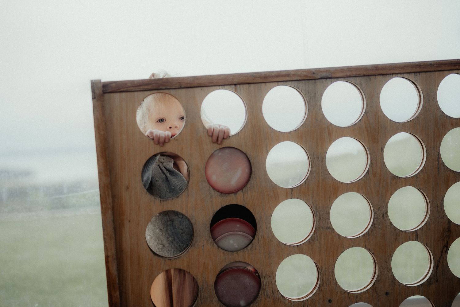 Child peeks through the holes of a giant wooden Connect Four game set up outdoors at a wedding.
