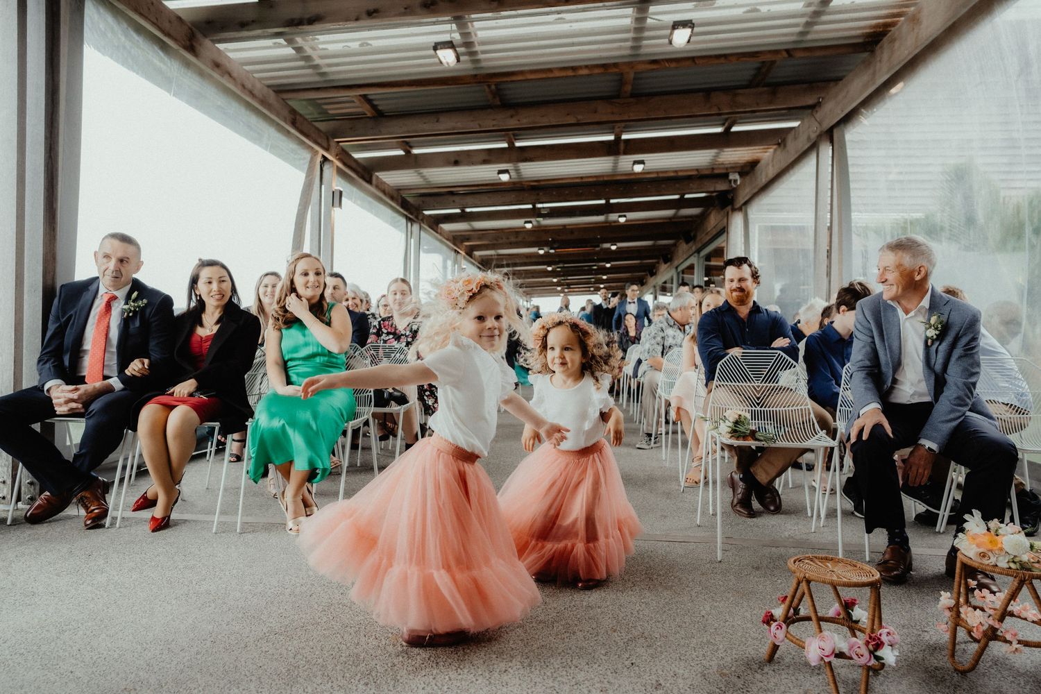 Two flower girls in pink tulle skirts walk down the aisle of a wedding ceremony as seated guests smile and watch.
