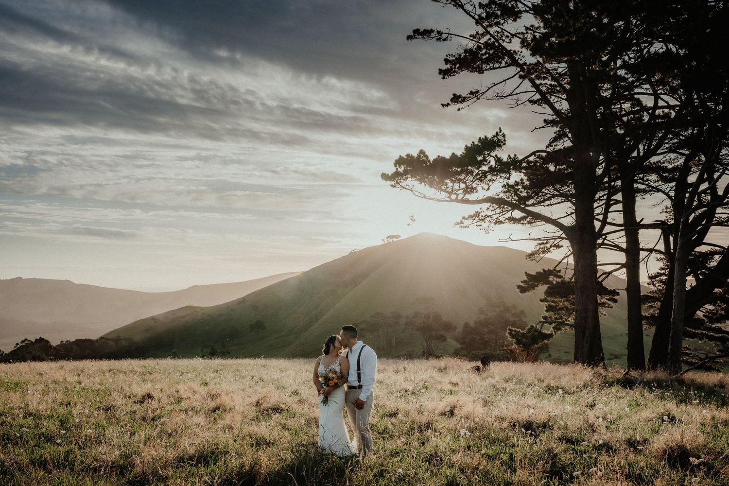 Bride and groom stand together on a grassy hill at sunset with dramatic clouds and a pine tree.
