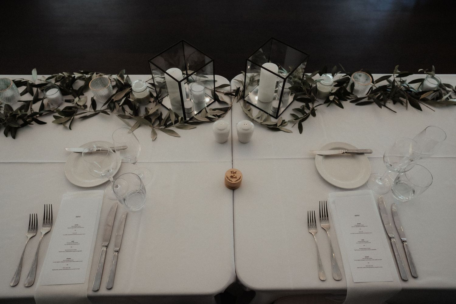 An elegant wedding reception table features white linens, olive branch garland, glass candle lanterns, menus and polished silverware.