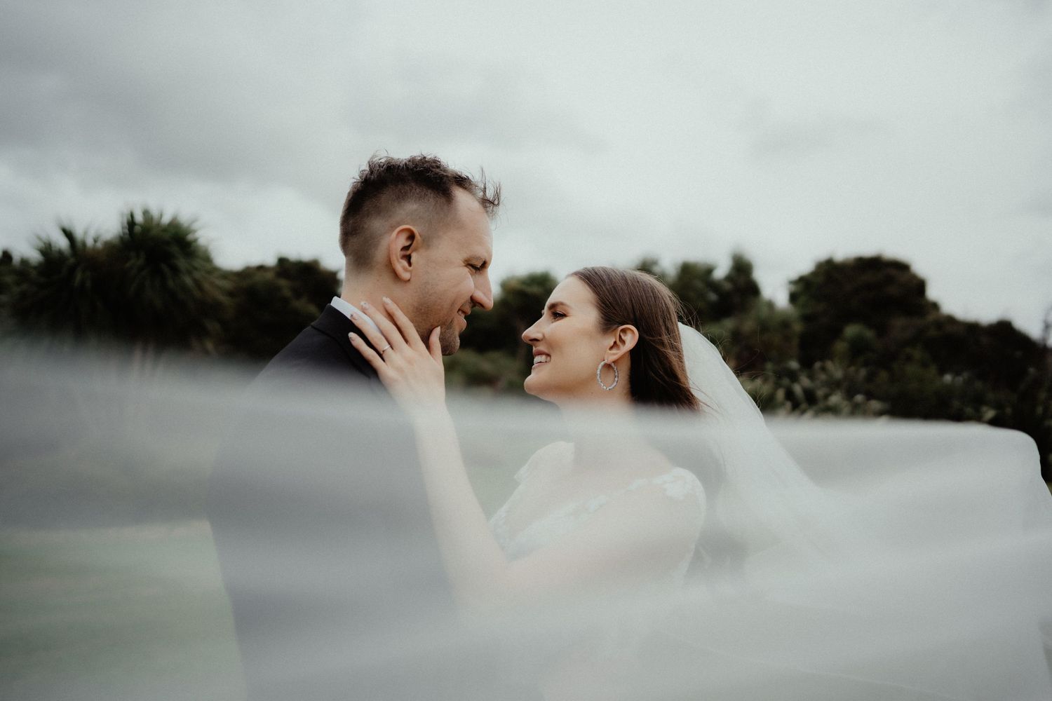 Happy bride touches her groom's face as they laugh together behind a soft sheer veil with trees in the background.