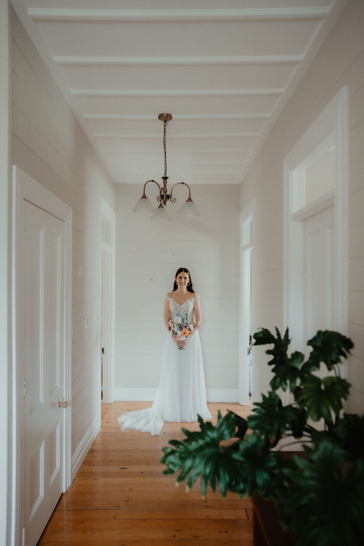 Bride in a lace wedding gown holding a bouquet stands in a bright hallway with a vintage chandelier and timber floors.