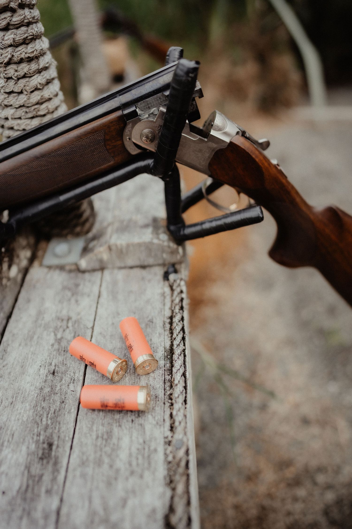 Close-up of an open double-barrel shotgun resting on a weathered wooden rail beside three orange shotgun shells.