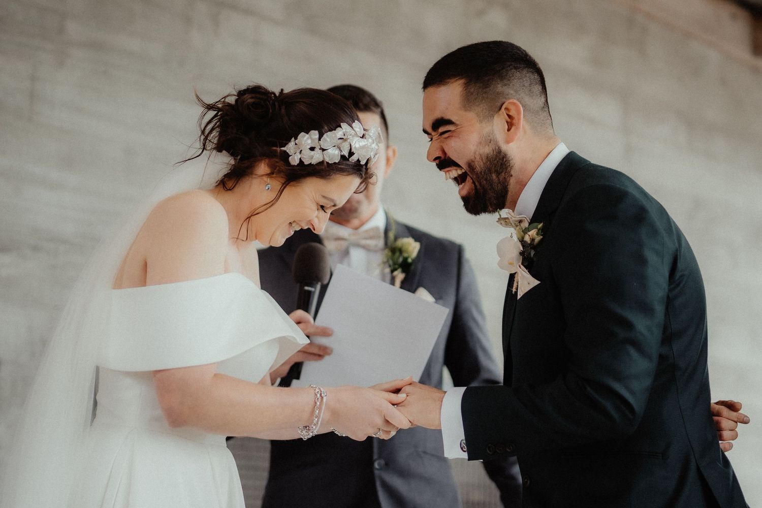 Laughing groom holds hands with the bride as she reads her vows from a paper during their wedding ceremony.