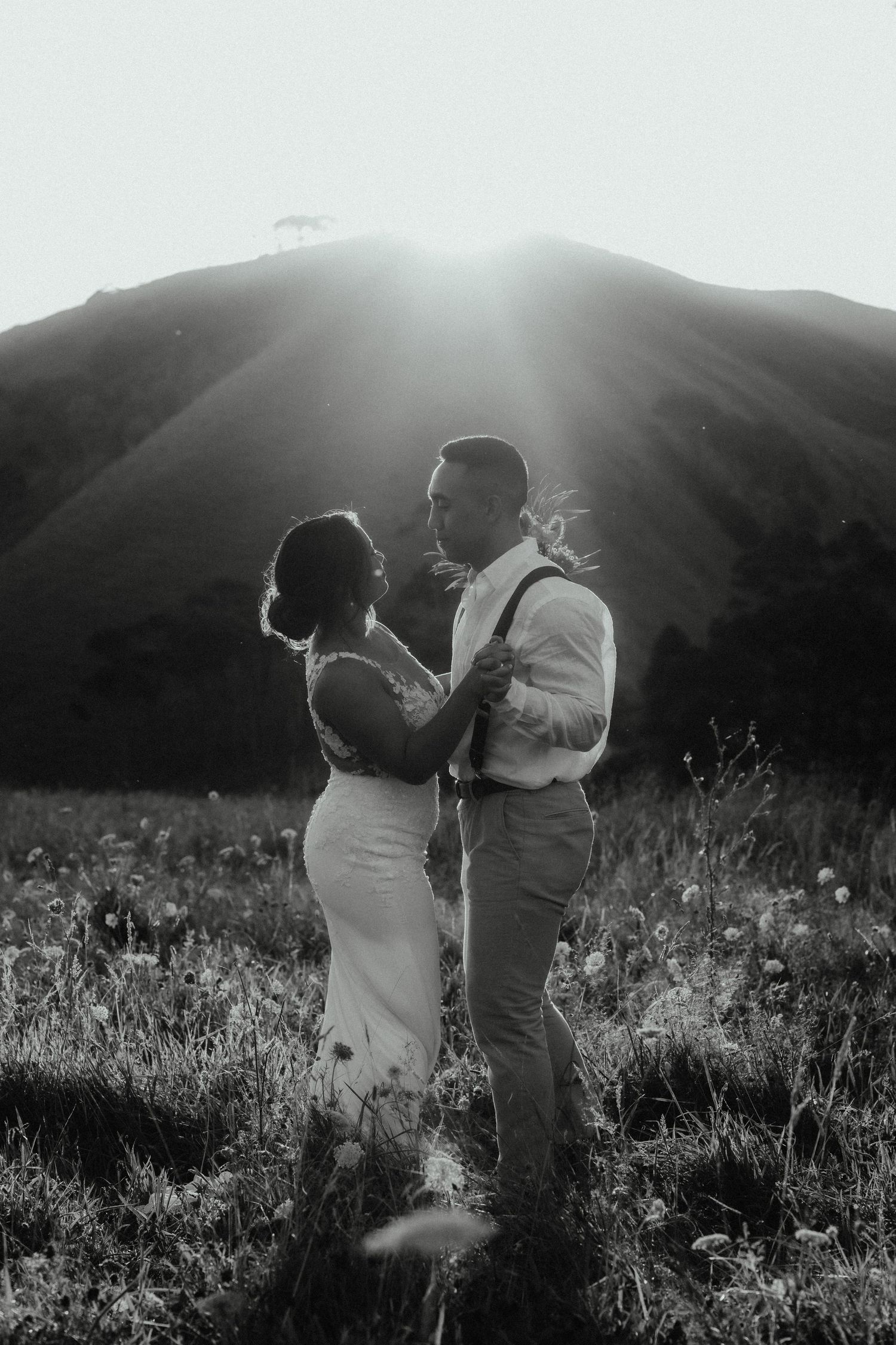 Black and white image of a couple dancing in a wildflower field as sunlight streams over a hill behind them.