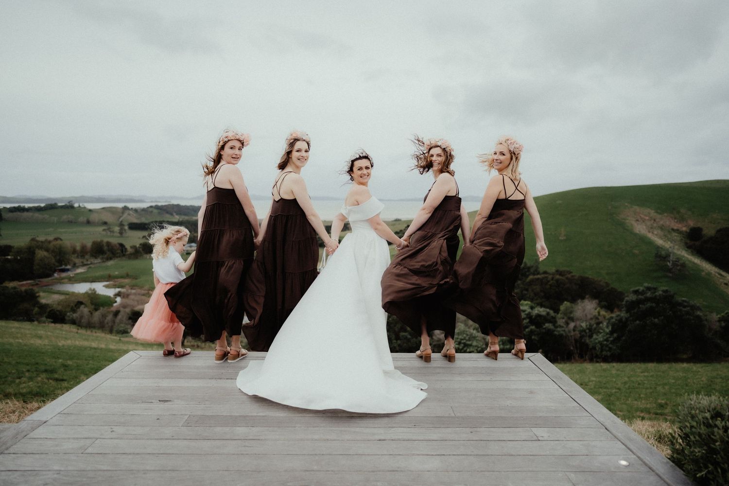 A bride in a white gown stands on a wooden deck with four bridesmaids in brown dresses and a flower girl overlooking green hills.