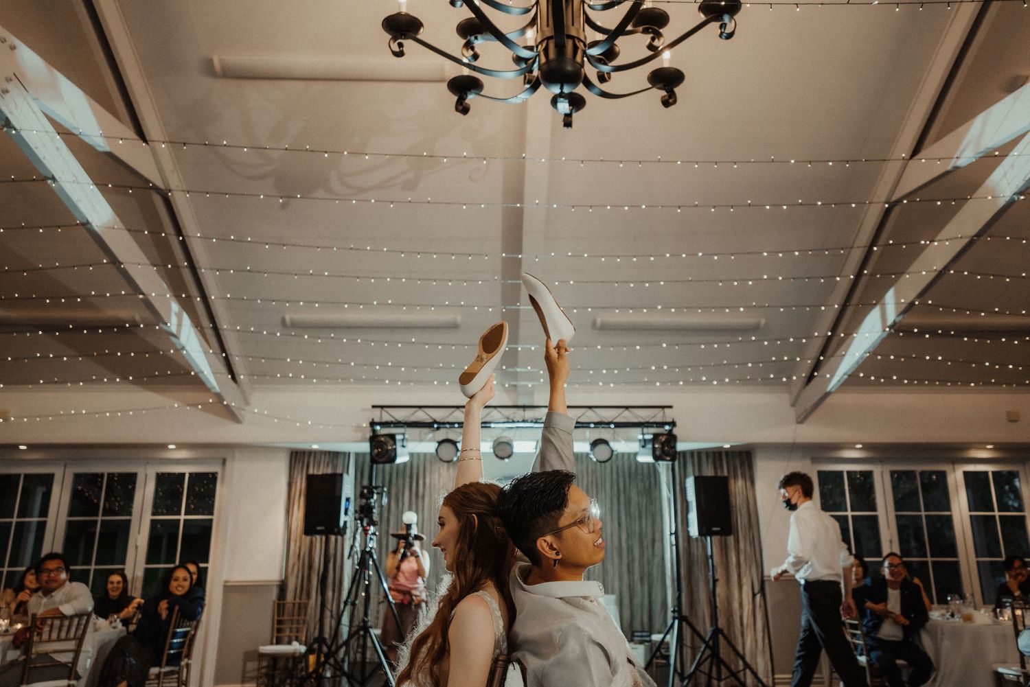 Wedding guests dance under a chandelier and string lights at the reception as a couple raises their arms in celebration.