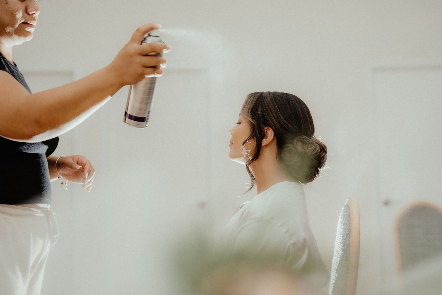 A hairstylist sprays hairspray onto a bride's updo as she sits in a bright room preparing for her wedding.