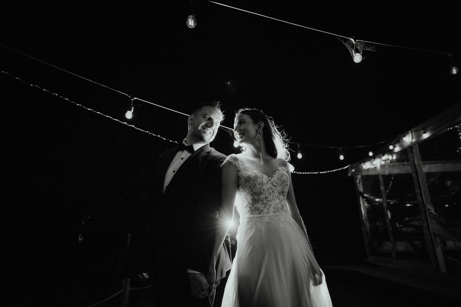 Black and white photo of a bride and groom sharing a romantic moment at night beneath glowing string lights at their wedding.