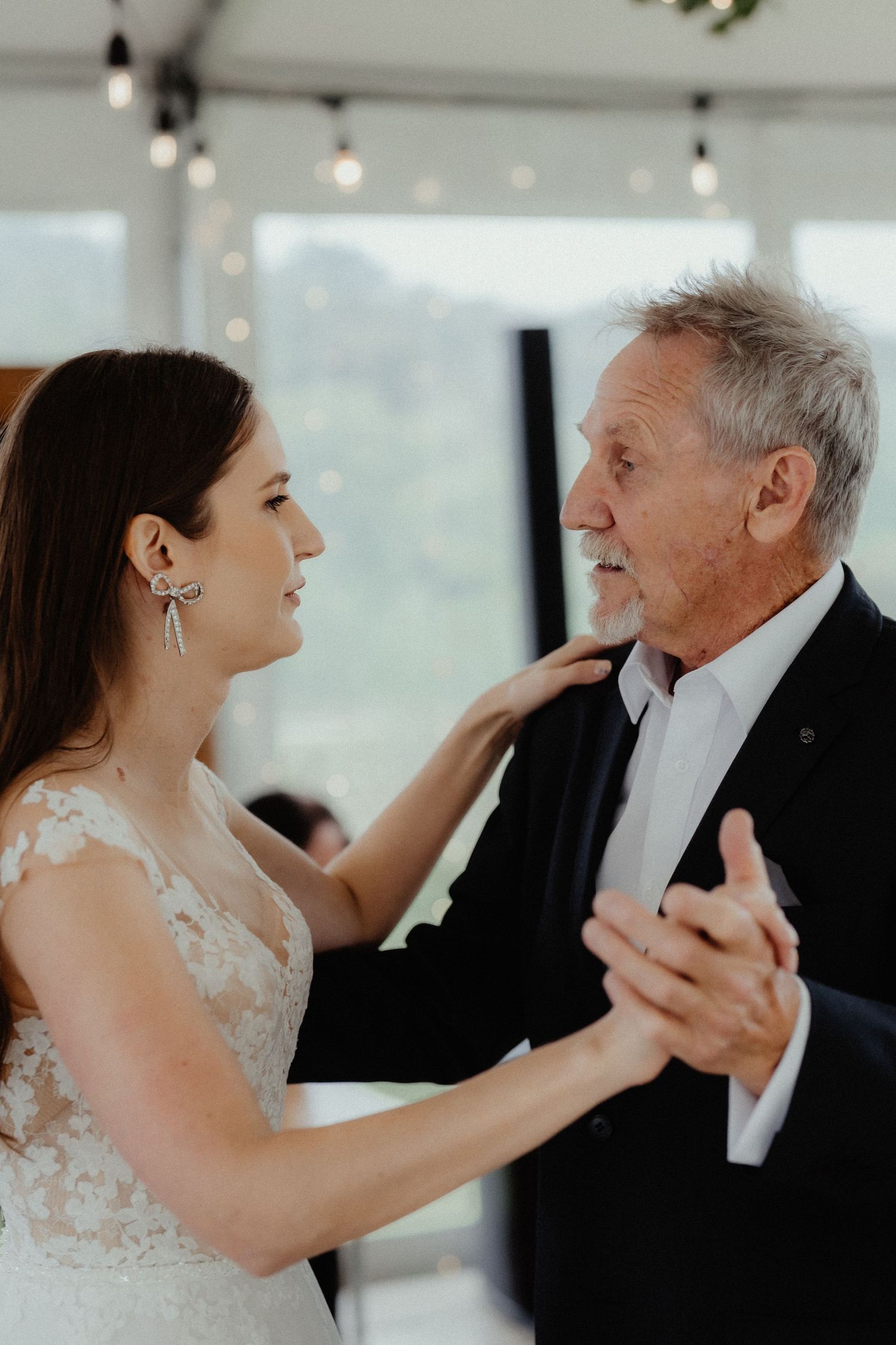 A bride in a lace gown and sparkling earrings shares a father-daughter dance with a smiling gray-haired man in a dark suit.