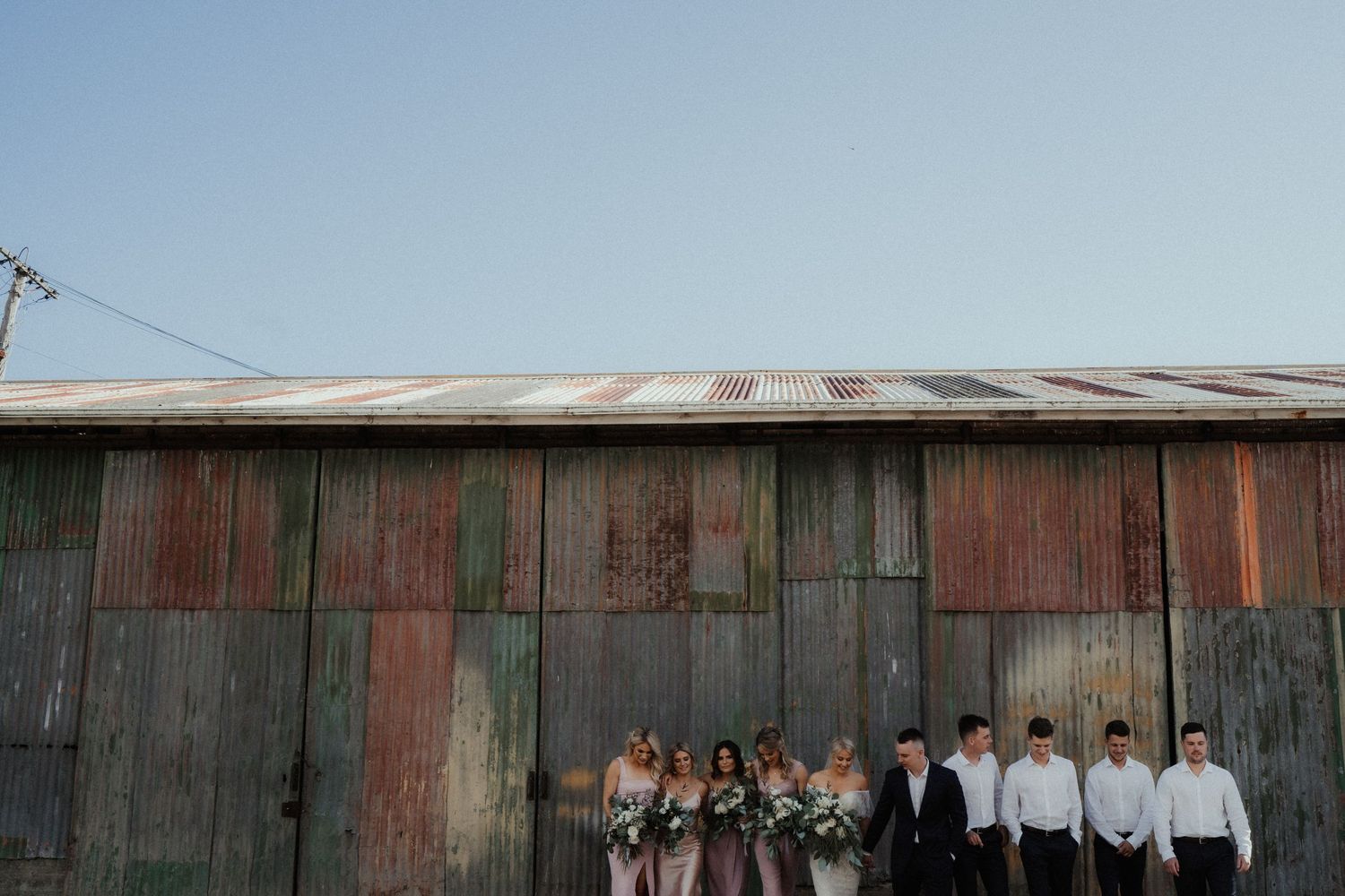 Bridal party of bridesmaids with bouquets and groomsmen in white shirts line up against a weathered rustic barn wall.