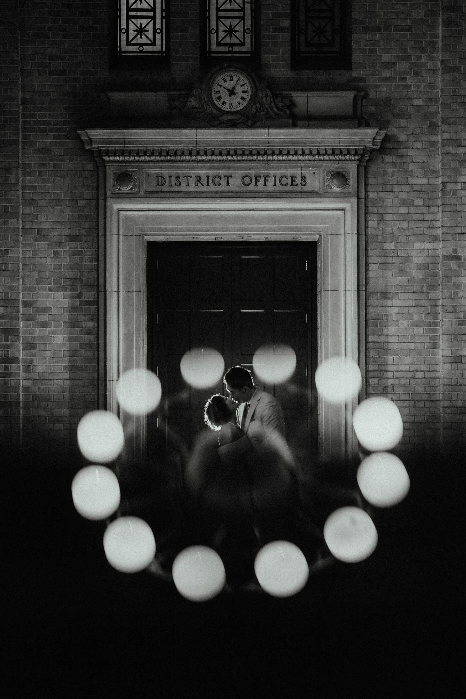 Black and white wedding photo of a couple kissing in front of a District Offices building with glowing circular lights around them.