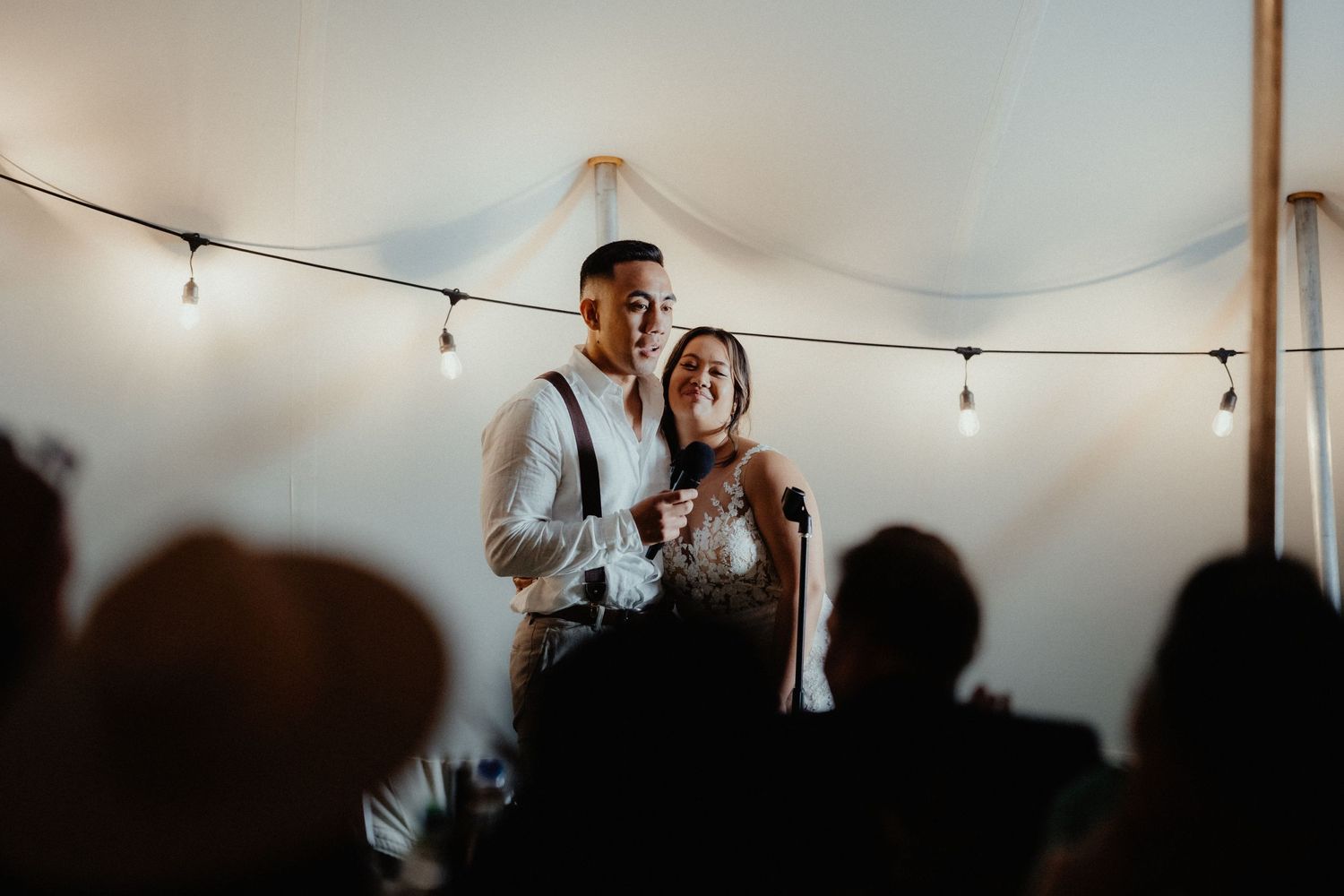 Smiling couple share a toast under string lights at their wedding reception as guests silhouette the foreground.