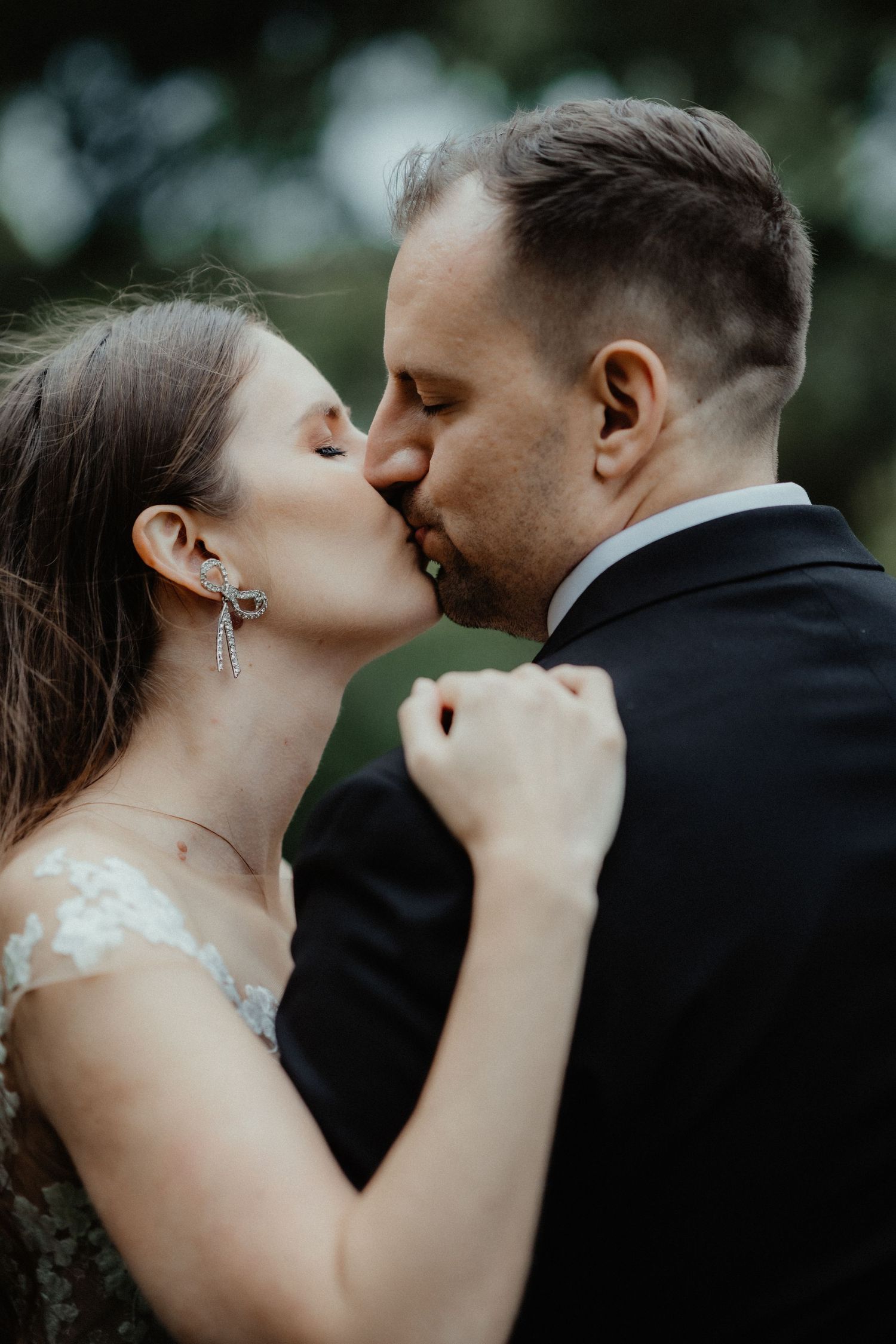 A close-up of a bride in lace and drop earrings kissing her groom in a dark suit during their wedding portraits.
