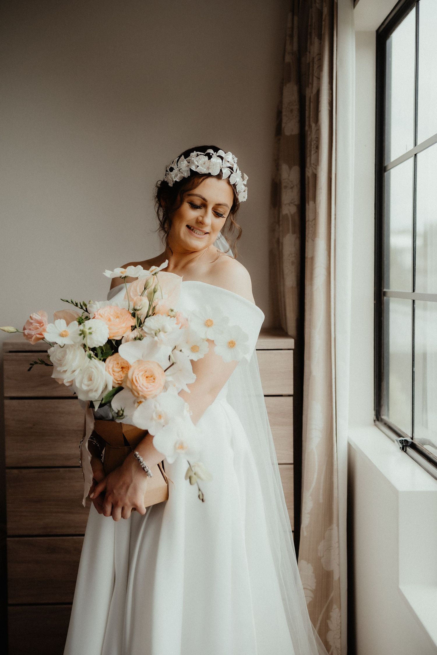 Bride in an off-shoulder white gown and floral headpiece holds a peach and white bouquet beside a large window.