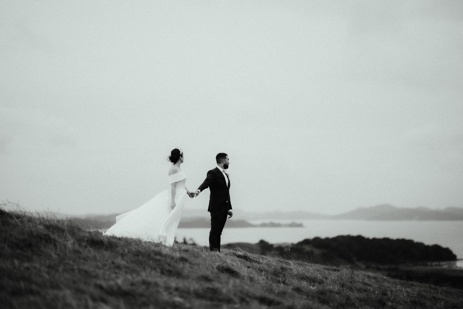 A black and white photo of a bride and groom holding hands on a grassy coastal hill as her veil blows in the wind.