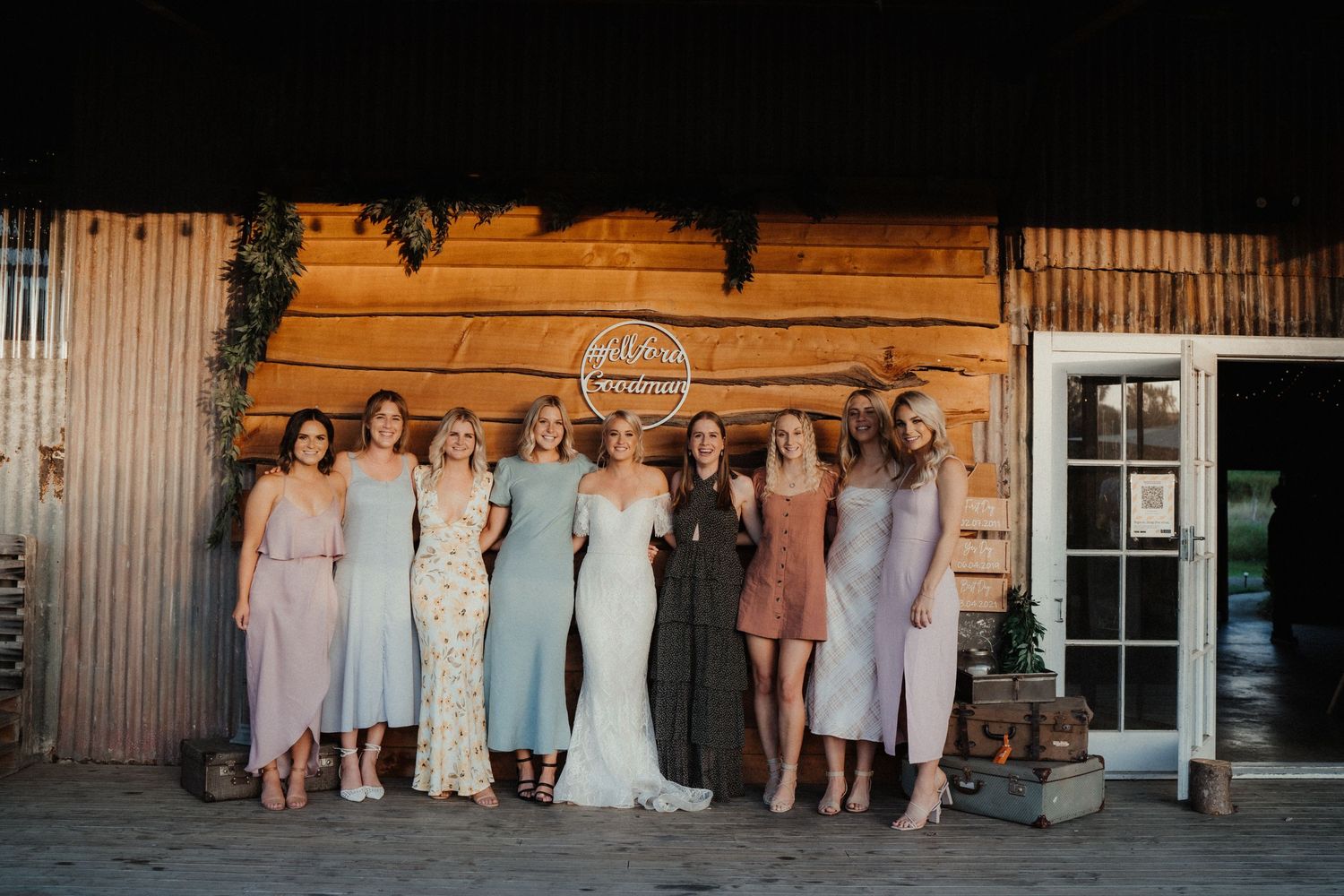 Bride stands with her bridesmaids in sage green and dusty pink dresses in front of a rustic timber barn wall.