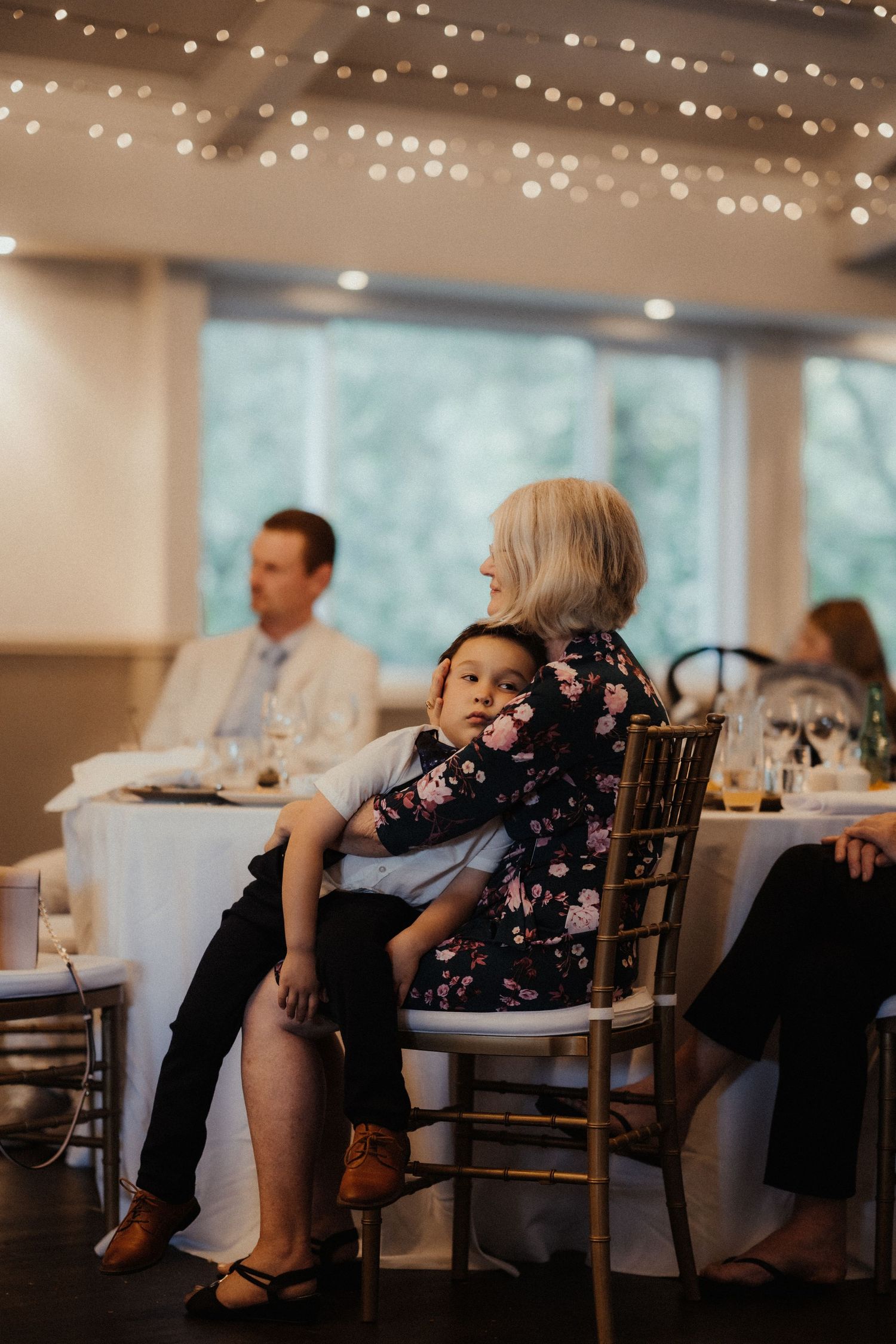 A young boy rests against a woman in a floral dress at a candlelit wedding reception table while guests dine nearby.