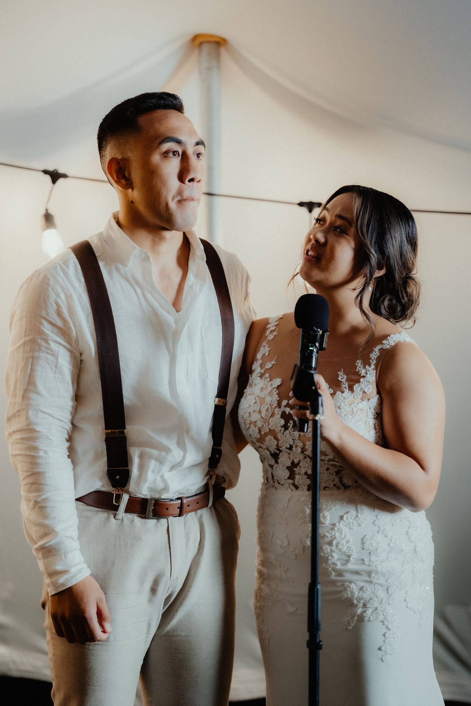 A bride and groom in suspenders and a lace gown stand at a microphone under a tent during their wedding reception.