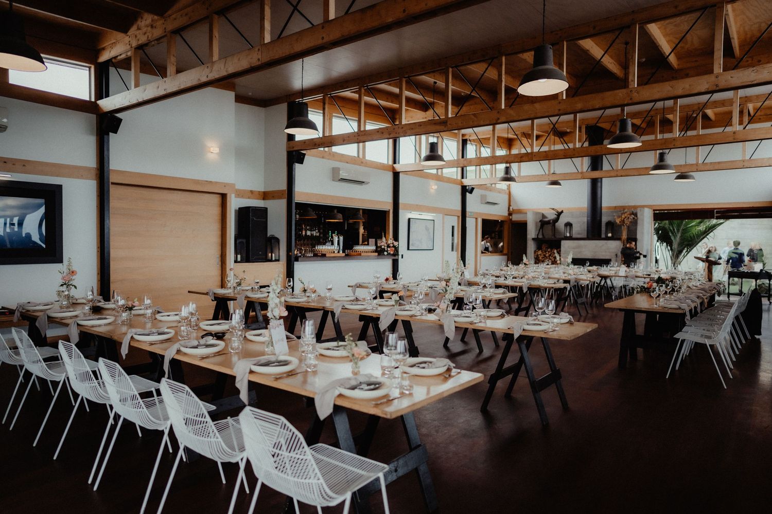Rustic wedding reception venue with long wooden tables, white chairs, pendant lights and exposed timber beams set for a celebration.