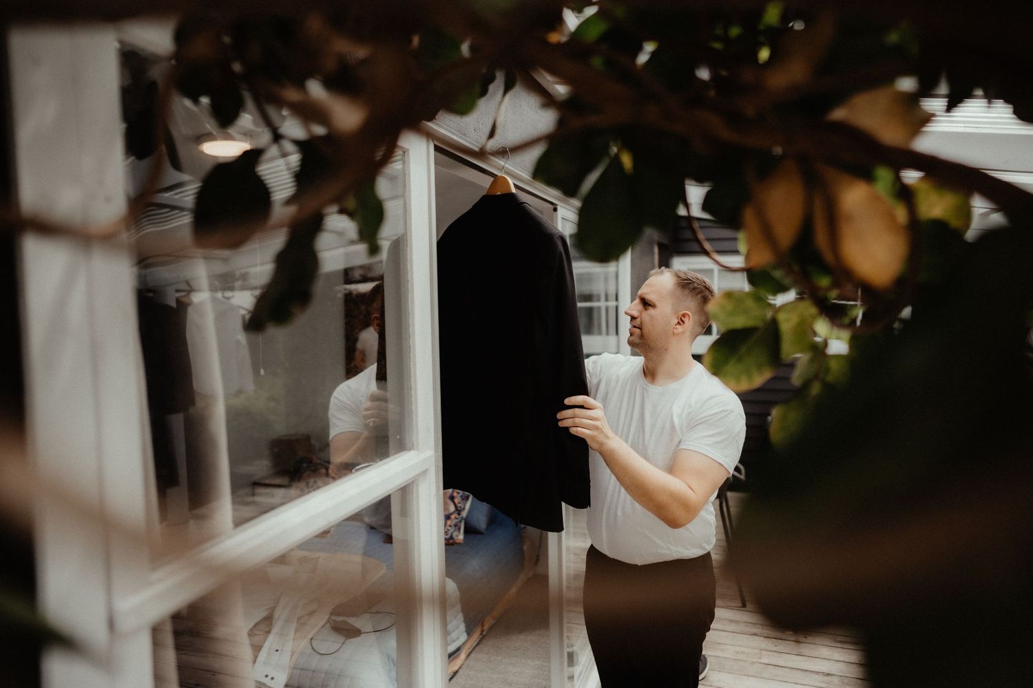A groom in a white t-shirt hangs his dark suit jacket on a rack near a window, framed by leaves, while getting ready for his wedding.