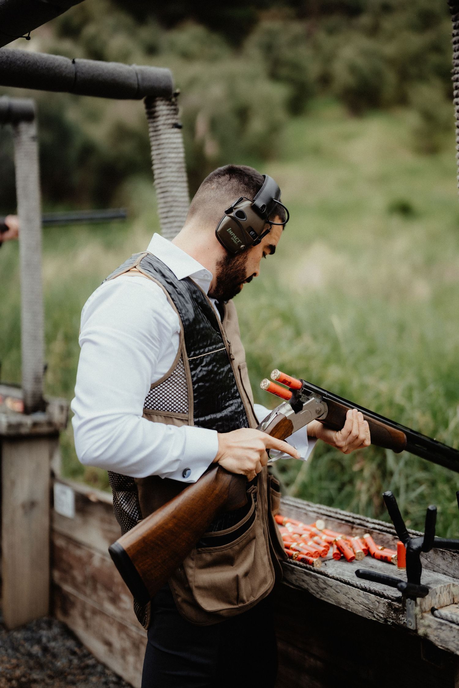 Groomsman wearing ear protection and a shooting vest loads a shotgun beside a table of red shells at an outdoor clay shooting range.