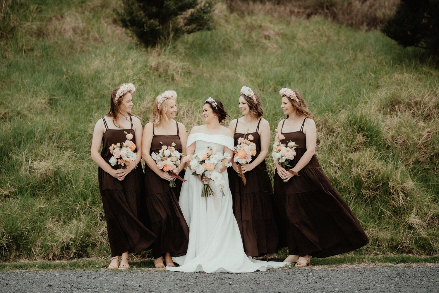 Bride in a white gown stands with four bridesmaids in brown dresses holding bouquets on a grassy hillside.