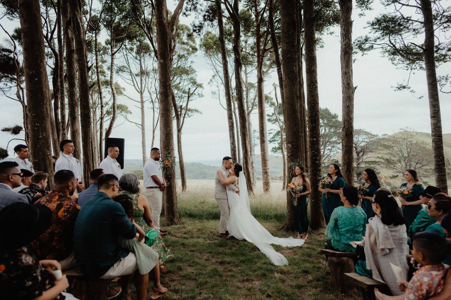 Outdoor wedding ceremony under tall trees as the couple exchange vows in front of seated guests.
