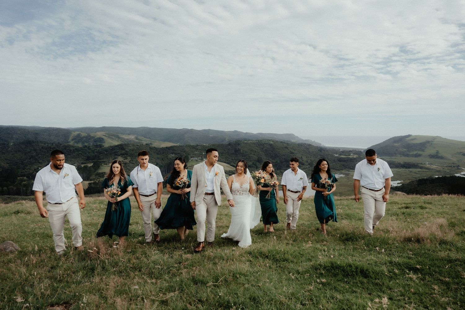 Bridal party in teal dresses and white shirts walk together with the bride and groom across a grassy hilltop on a cloudy day.