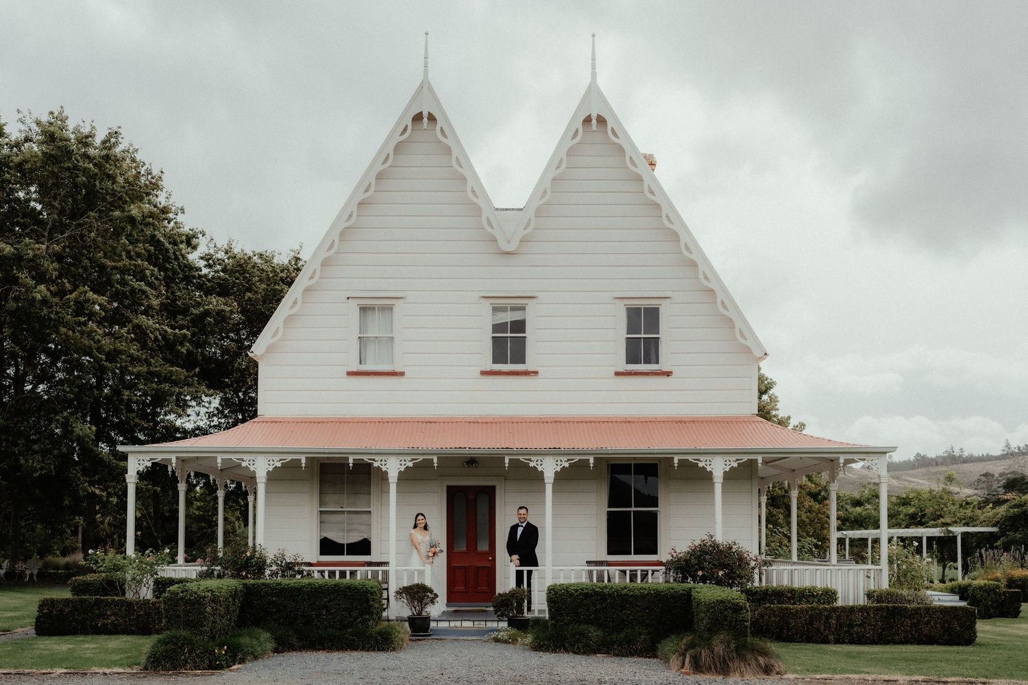 Bride and groom stand on the front steps of a white Victorian-style country house with a red door and veranda.