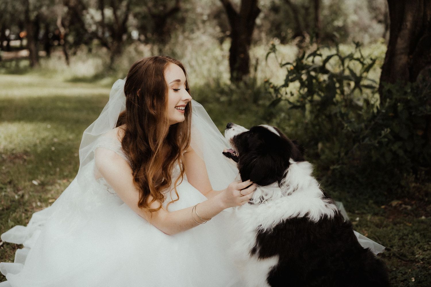 A smiling bride in a flowing white wedding gown gently pets a black and white dog in a leafy outdoor setting.