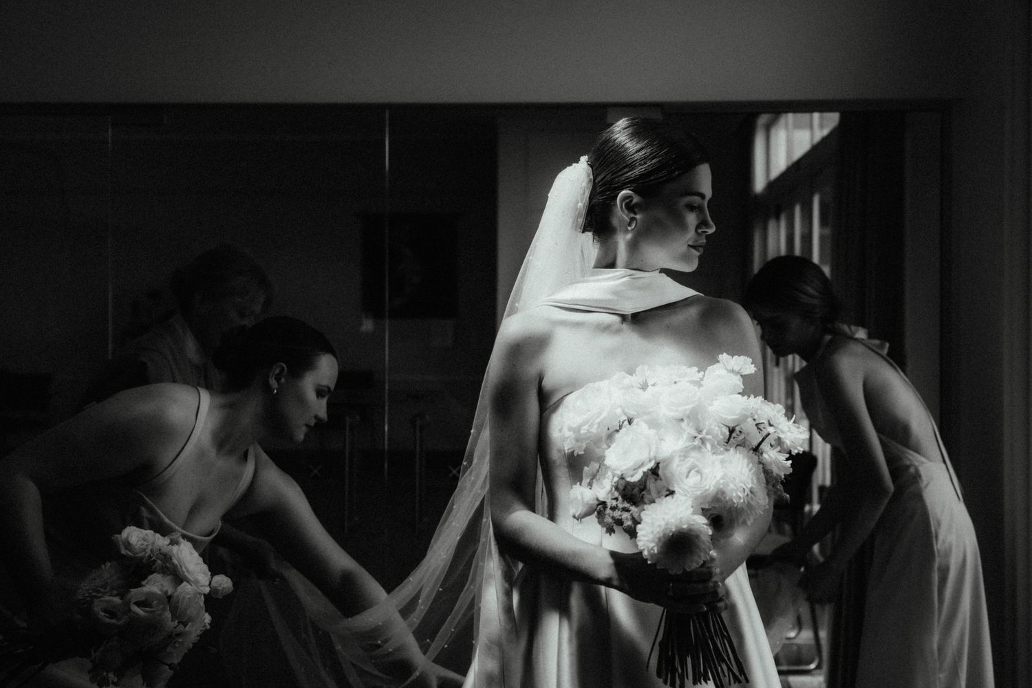 Black and white photo of a bride in a strapless gown and veil holding a white bouquet while bridesmaids help prepare in the background.