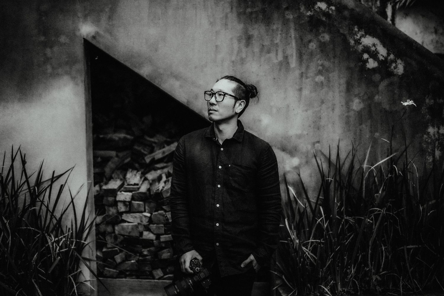Black and white portrait of a man with glasses and a topknot standing beside a stone wall and tall grass.