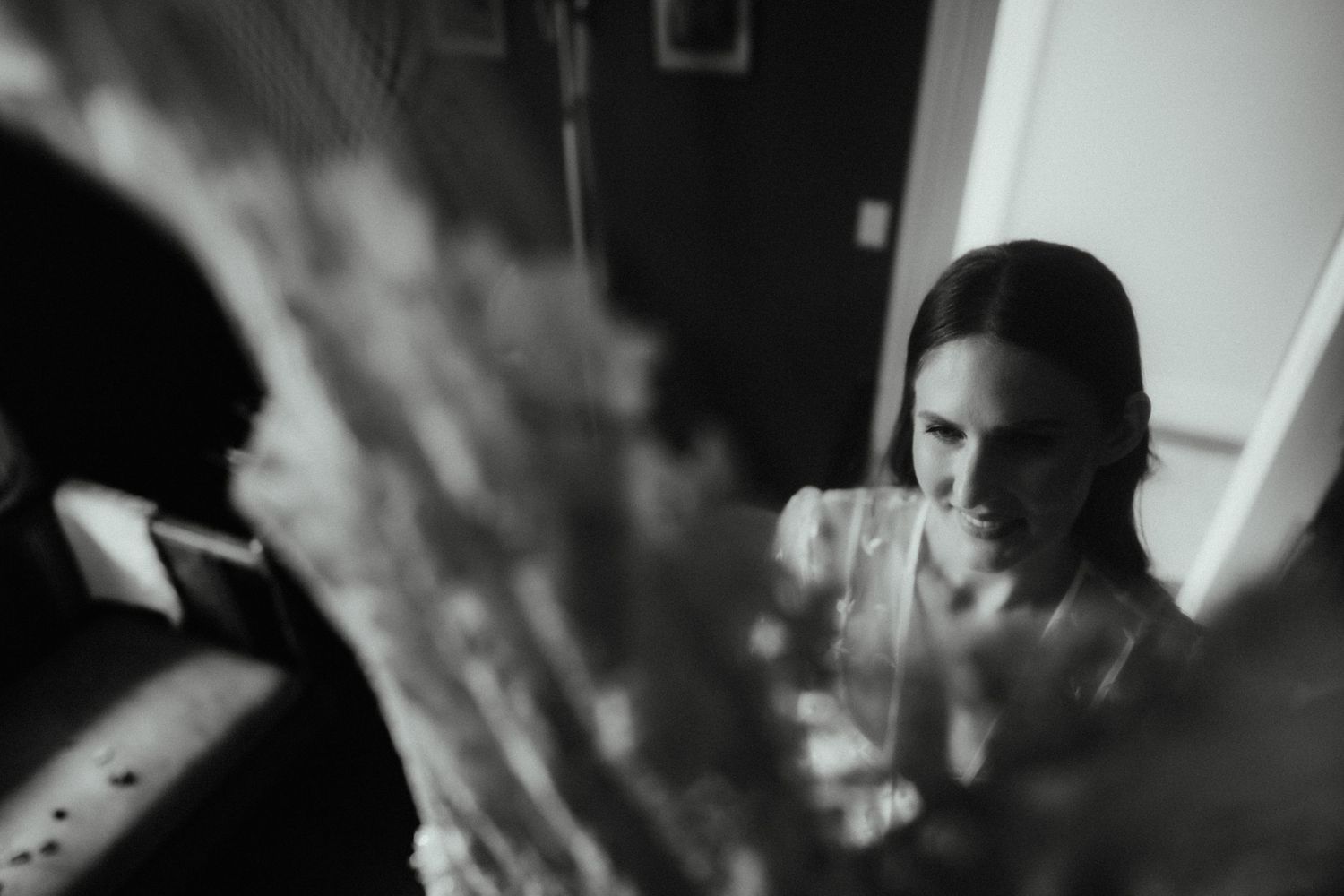 Black and white candid of a smiling woman looking at flowers in soft focus during wedding preparations.