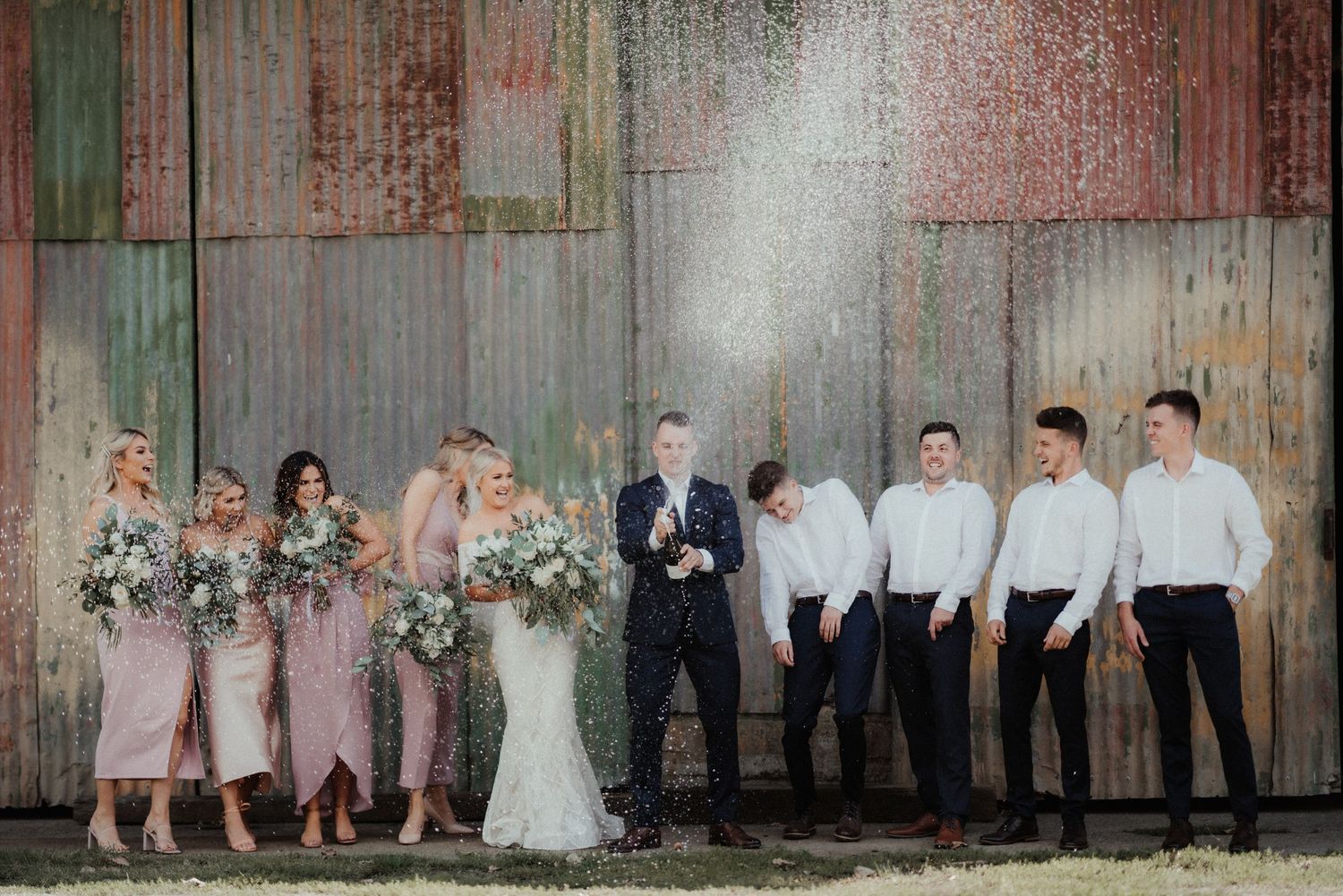 Groom sprays champagne in front of a rustic barn wall while the bride, bridesmaids with bouquets and groomsmen laugh and celebrate.