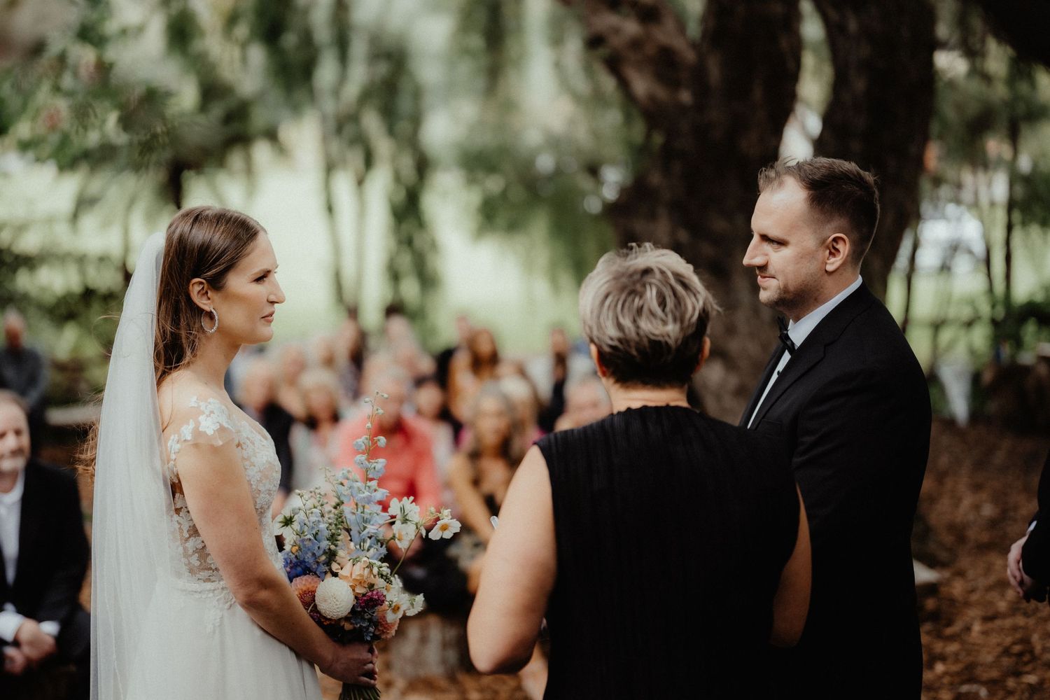 Bride in a lace gown and veil holds a colorful bouquet while facing the groom during an outdoor wedding ceremony with guests watching.
