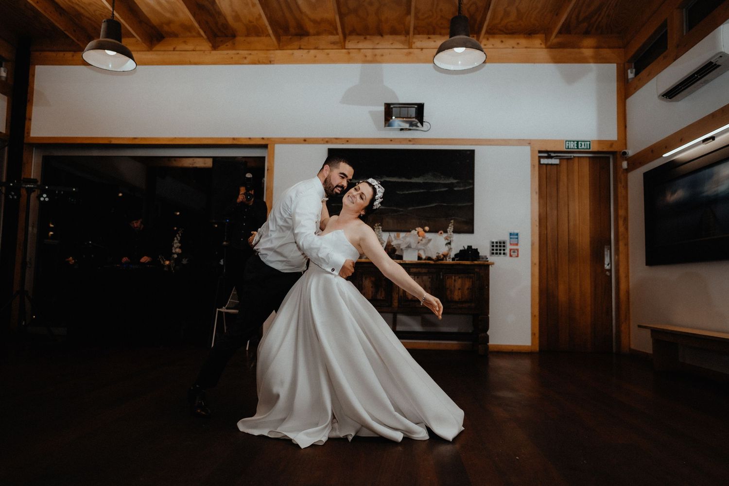 Groom dips the bride in a romantic dance pose on a wooden floor inside a rustic wedding venue with pendant lights.