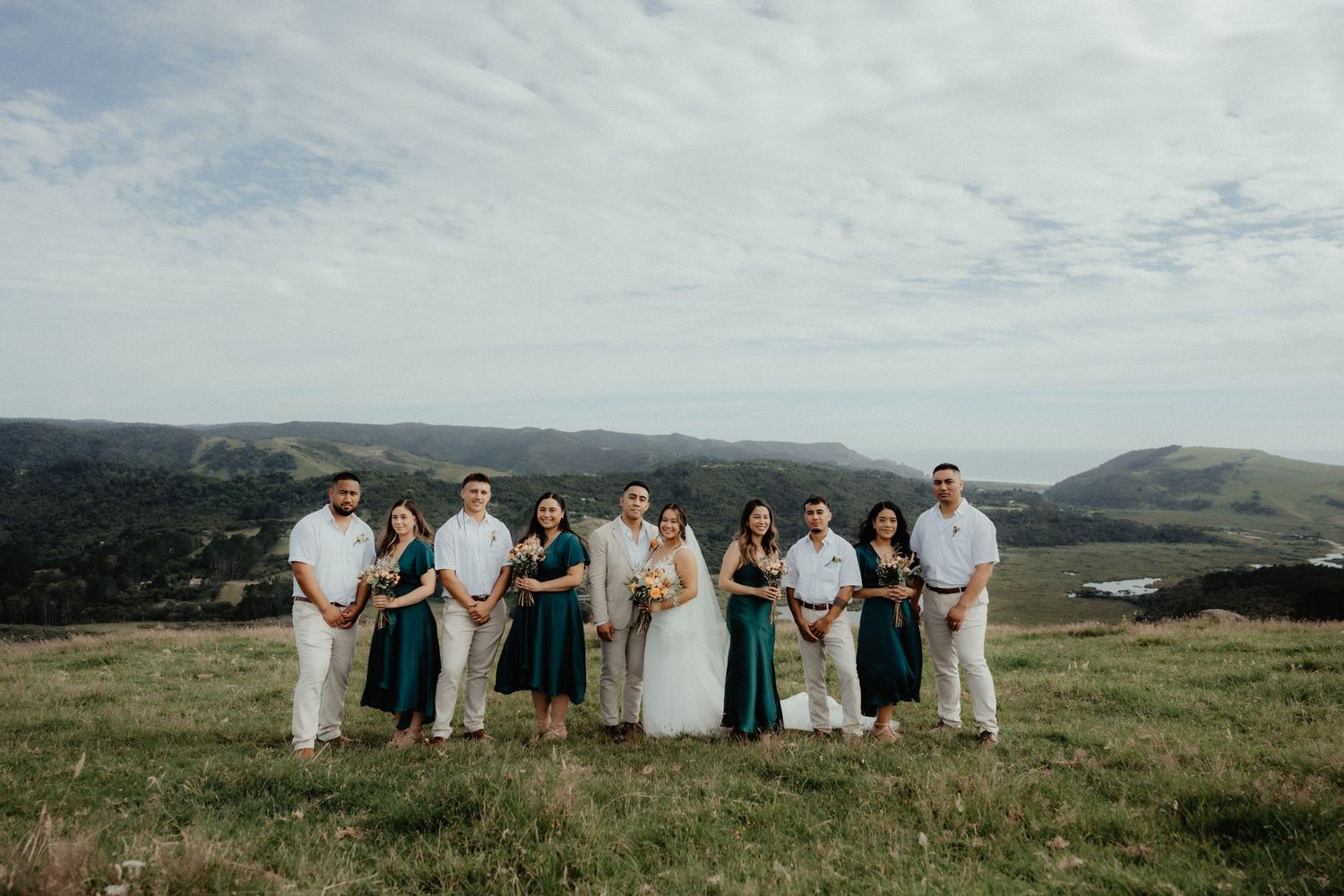 Wedding party of bride, groom, groomsmen and bridesmaids in teal dresses poses together on a grassy hilltop under a cloudy sky.