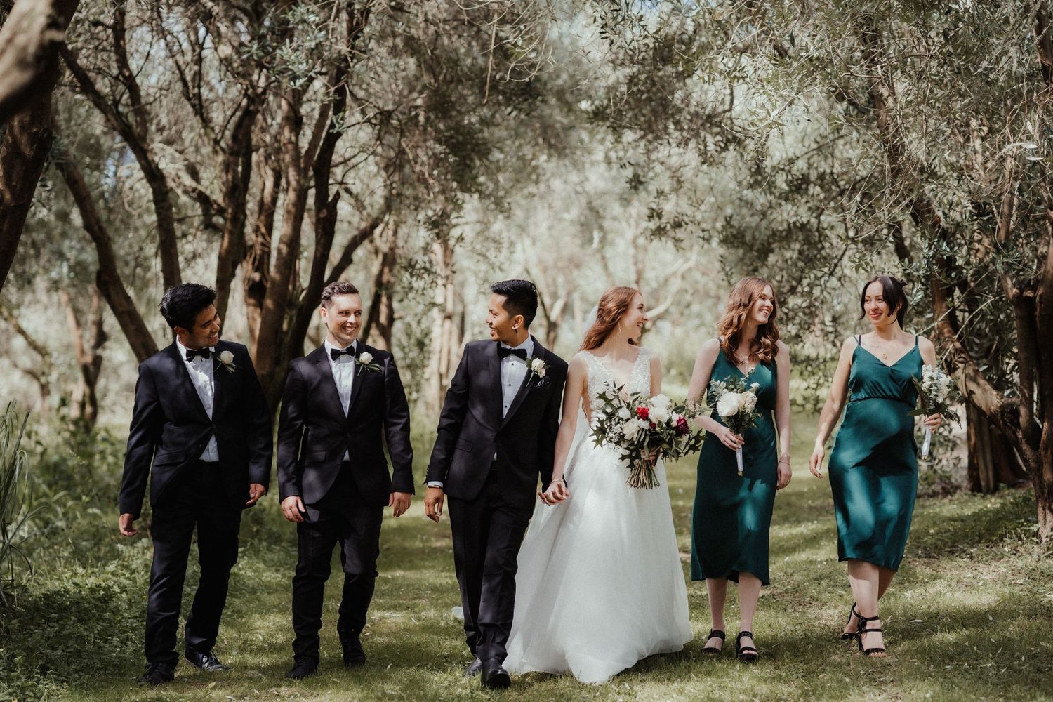 Three groomsmen in dark suits walk arm in arm with a bride and two bridesmaids in green dresses through a sunlit olive grove.
