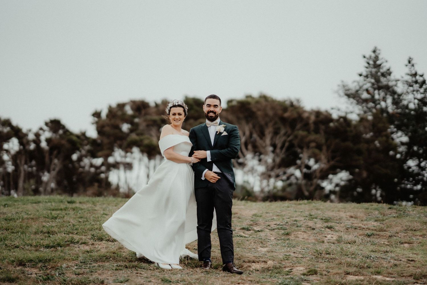 Smiling bride and bearded groom in a dark suit embrace on a grassy hilltop with windswept trees behind them.
