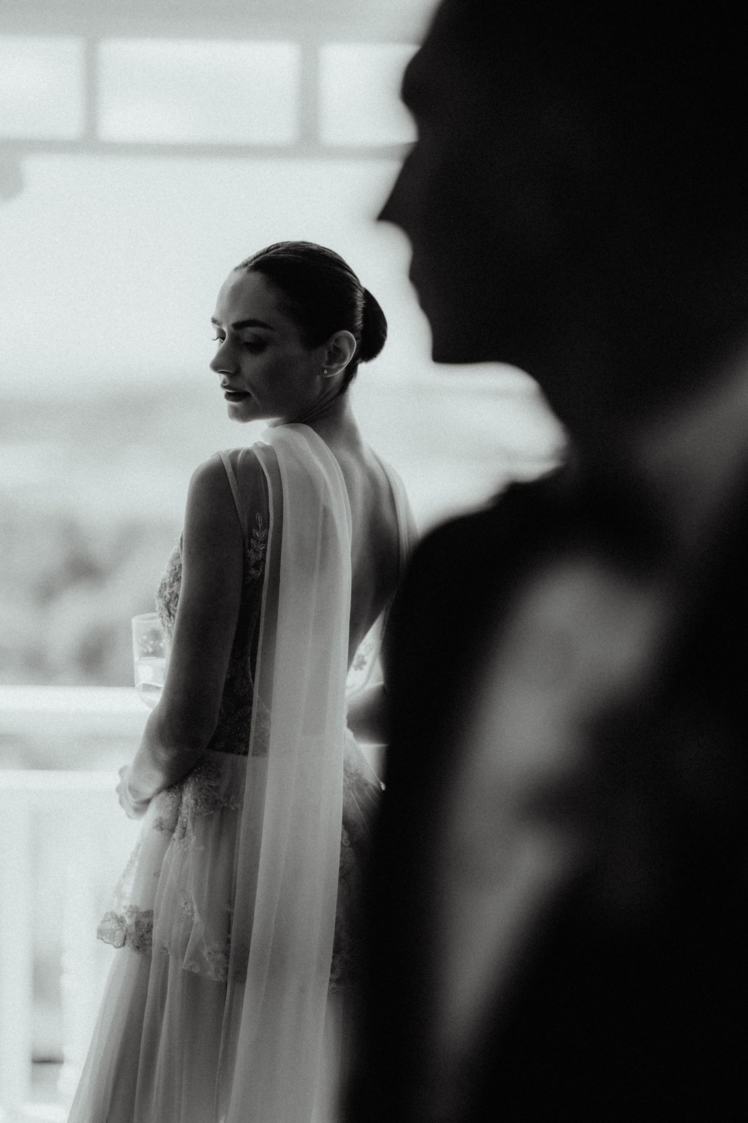 Black and white photo of a bride in a lace gown with a veil looking down, with the groom's silhouette blurred in the foreground.