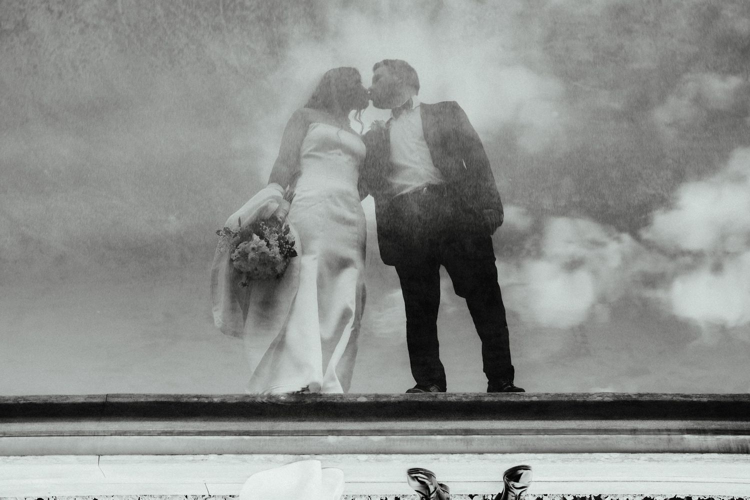 Bride and groom kiss on a stone ledge under a cloudy sky in a moody black and white wedding photograph.